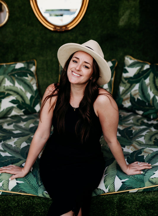 headshot of Ashlee Kinsella of Tiny Human Print Co sitting on greenery couch in black dress and white hat